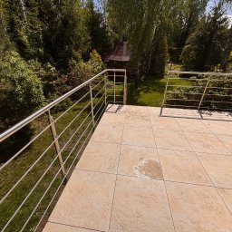 Adrian Durak - Beżowy taras z metalową balustradą, widok na zielony ogród z altaną. Płytki tarasowe w ciepłym odcieniu, nowoczesne ogrodzenie ze stali nierdzewnej. Słoneczny dzień.