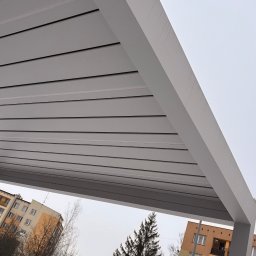 Adrian Durak - Nowoczesna, szara pergola z widokiem na budynki mieszkalne i drzewa w zimowej scenerii. Ujęcie z dołu, podkreślające minimalistyczny design konstrukcji.