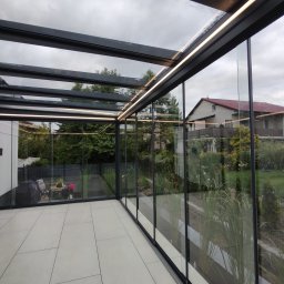 Adrian Durak - Nowoczesna, przeszklona konstrukcja z oświetleniem LED, tworząca osłoniętą przestrzeń z widokiem na ogród i dom. Minimalistyczny design, szklane ściany.