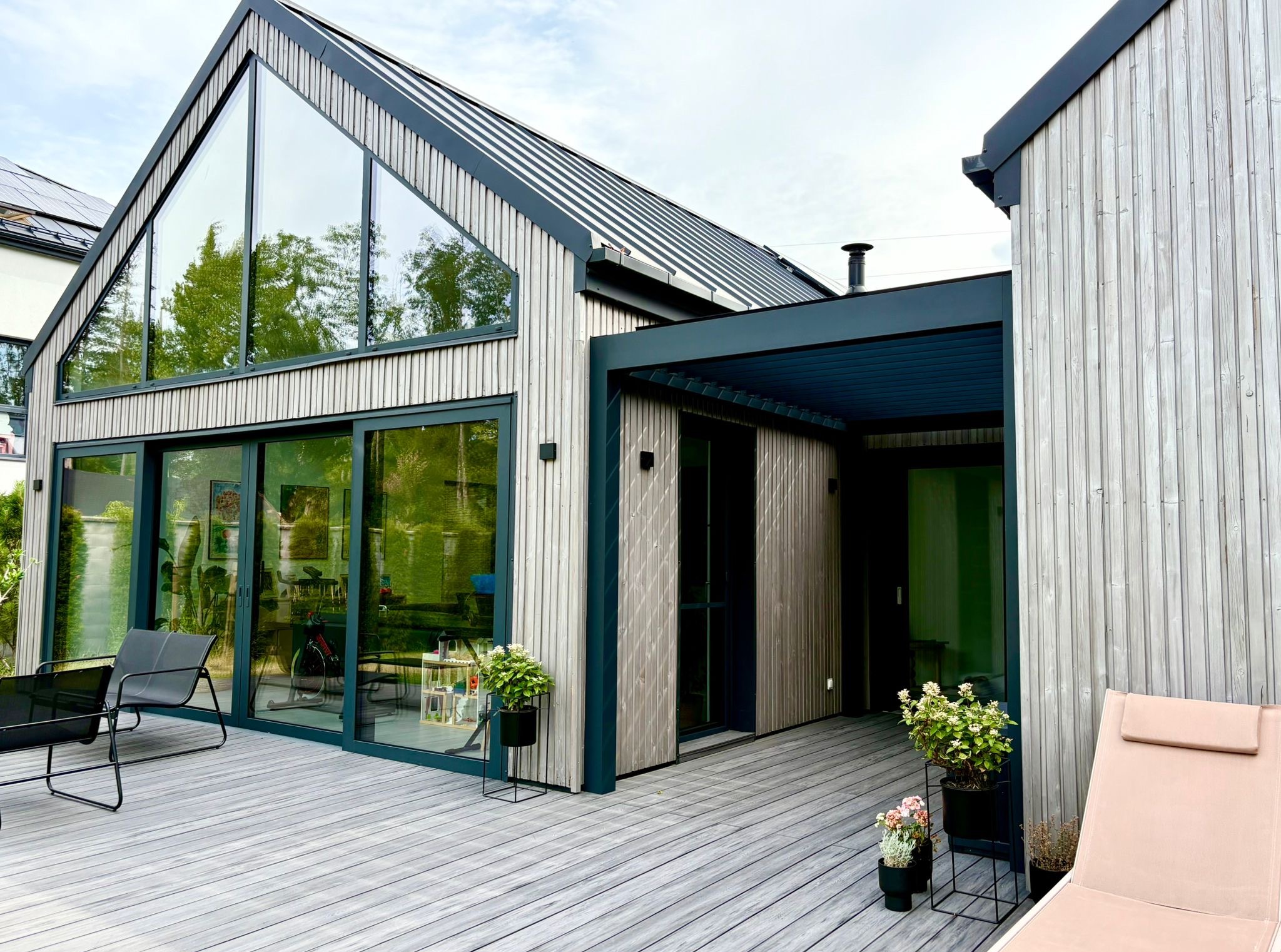 Nowoczesny dom z drewnianą elewacją, tarasem kompozytowym i pergolą. Widoczne meble ogrodowe i rośliny w donicach. Duże przeszklenia odbijają zieleń ogrodu.