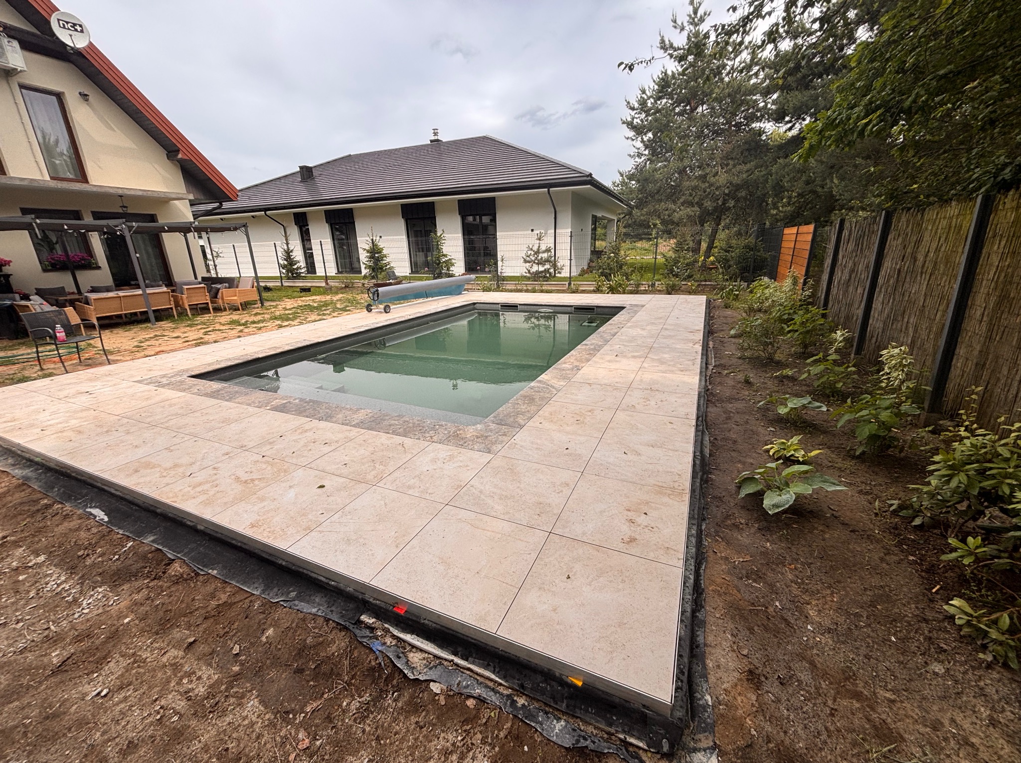 Nowoczesny taras z jasnych płyt wokół basenu, widok na dom z ciemnym dachem i ogrodzenie z bambusa. Ujęcie z poziomu gruntu, podkreślające detale wykończenia.