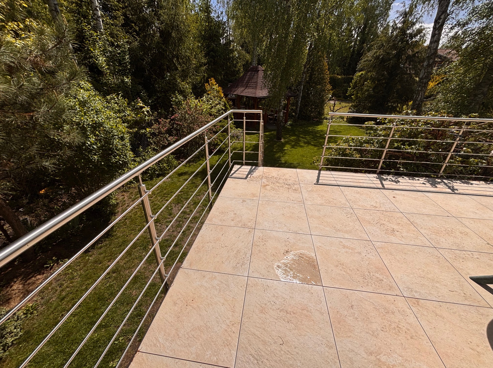 Beżowy taras z metalową balustradą, widok na zielony ogród z altaną. Płytki tarasowe w ciepłym odcieniu, nowoczesne ogrodzenie ze stali nierdzewnej. Słoneczny dzień.