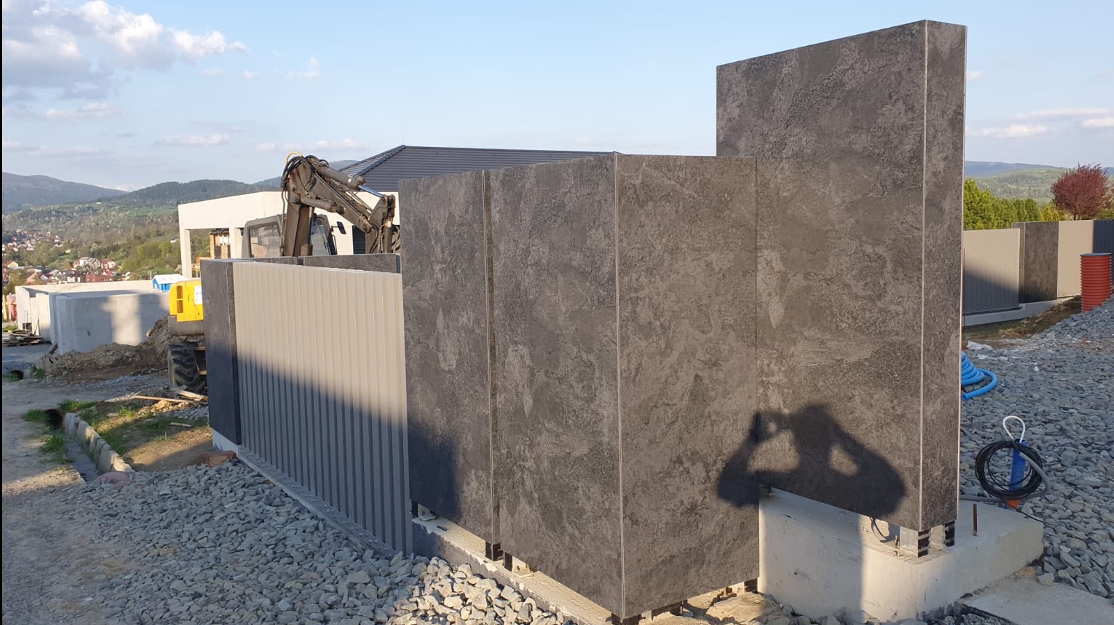 Montaż nowoczesnego ogrodzenia z paneli imitujących kamień, widoczne elementy konstrukcyjne i sprzęt budowlany w tle, krajobraz górski.