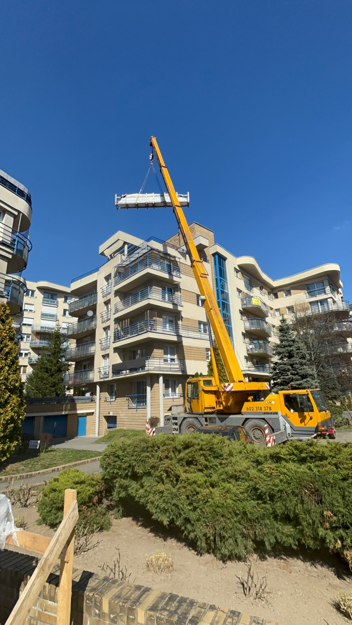 Żuraw budowlany podnosi element konstrukcyjny na tle budynku mieszkalnego z balkonami. Błękitne niebo w tle. Widoczne krzewy i fragment murku.