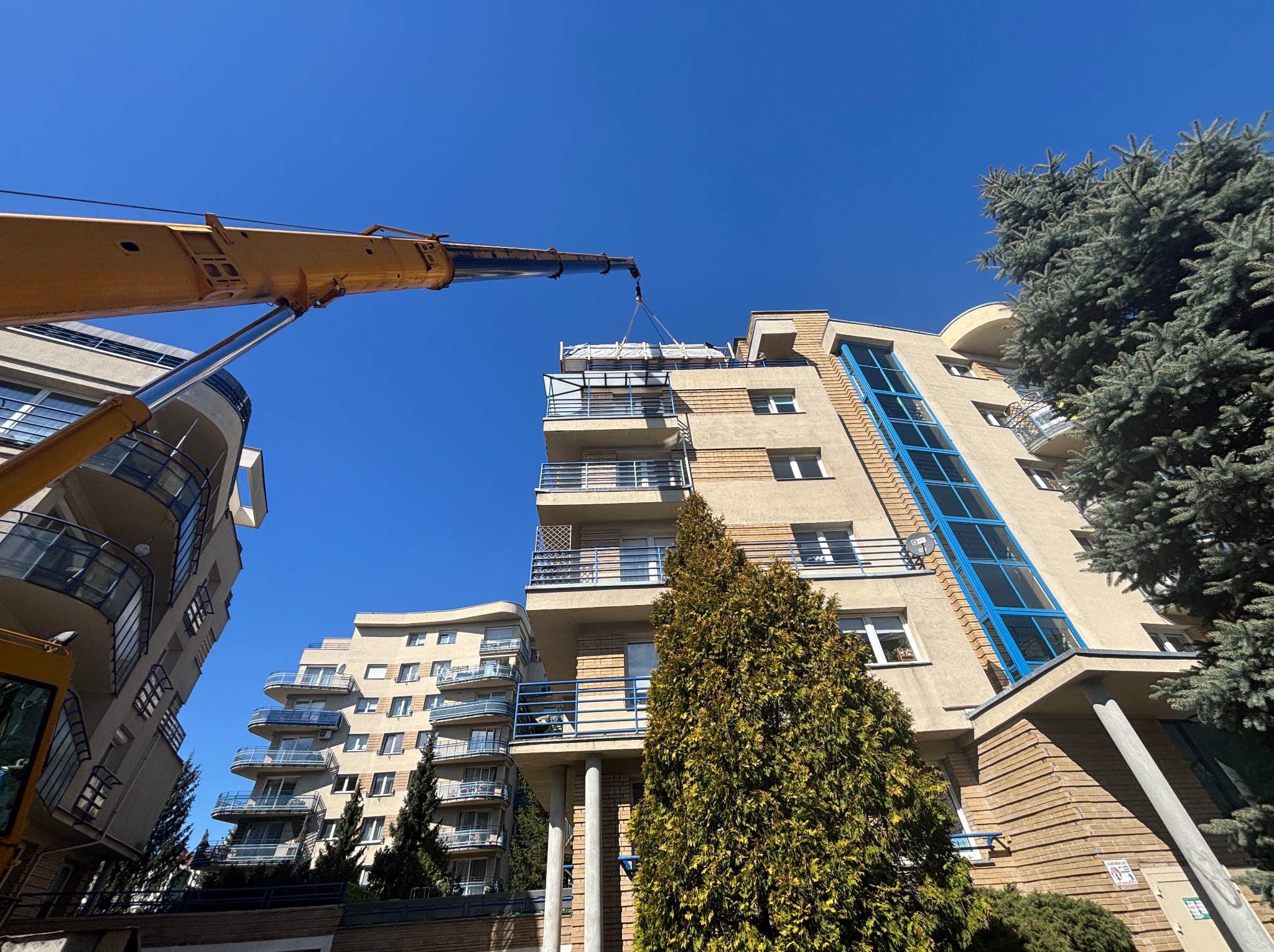 Montaż elementów na balkonie bloku mieszkalnego za pomocą dźwigu. Widok z dołu, błękitne niebo w tle. Prace na wysokościach, zabezpieczony ładunek.