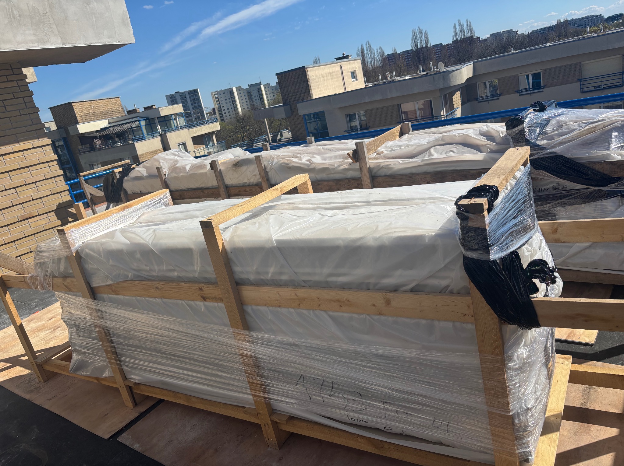 Paczki z elementami tarasu kompozytowego, zabezpieczone folią i drewnianą konstrukcją, czekają na montaż na tle miejskiego krajobrazu.