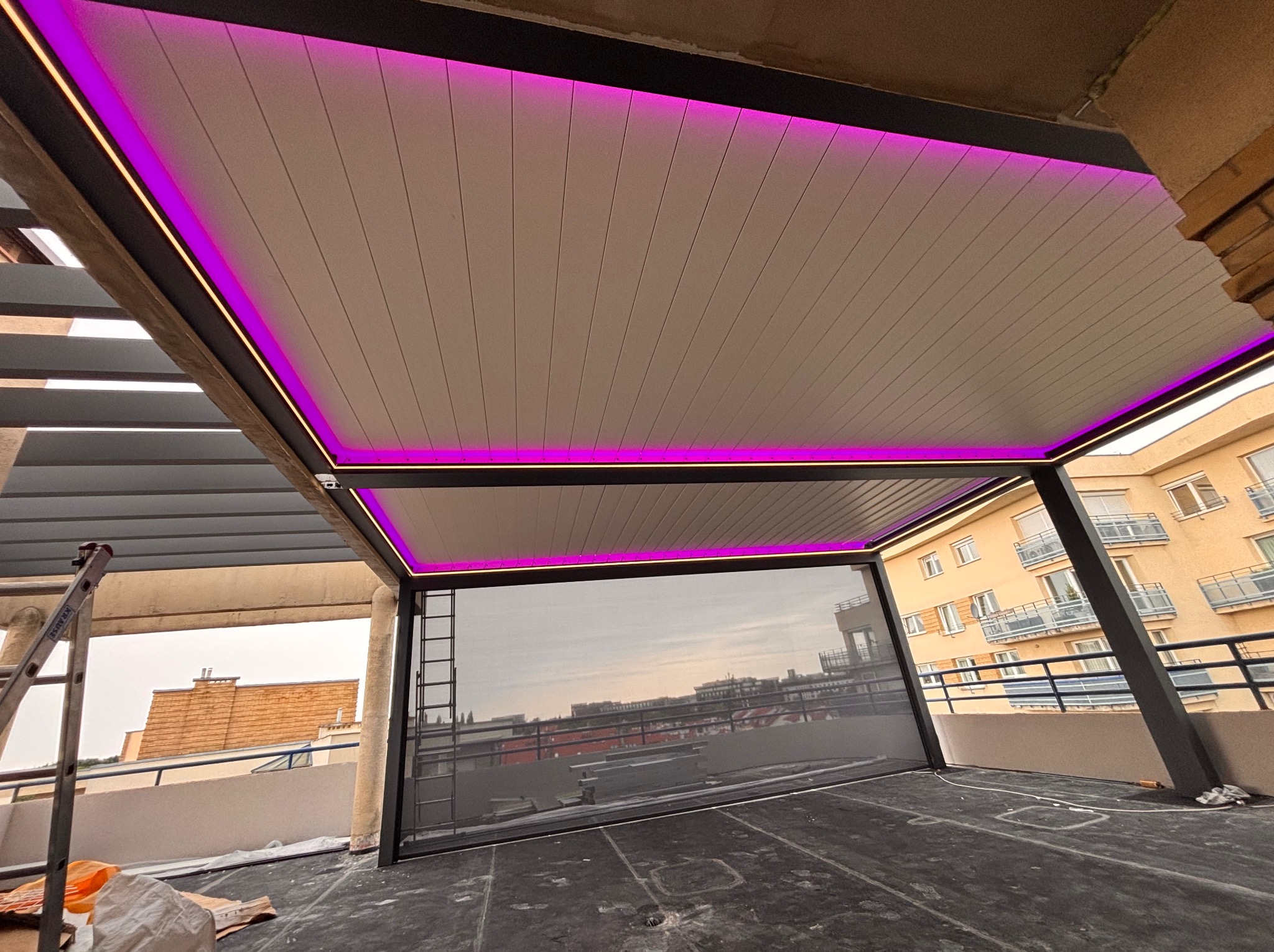 Nowoczesna pergola z metalową konstrukcją i oświetleniem LED w kolorze fioletowym, zamontowana na balkonie z widokiem na miasto. Widoczna drabina i elementy montażowe.