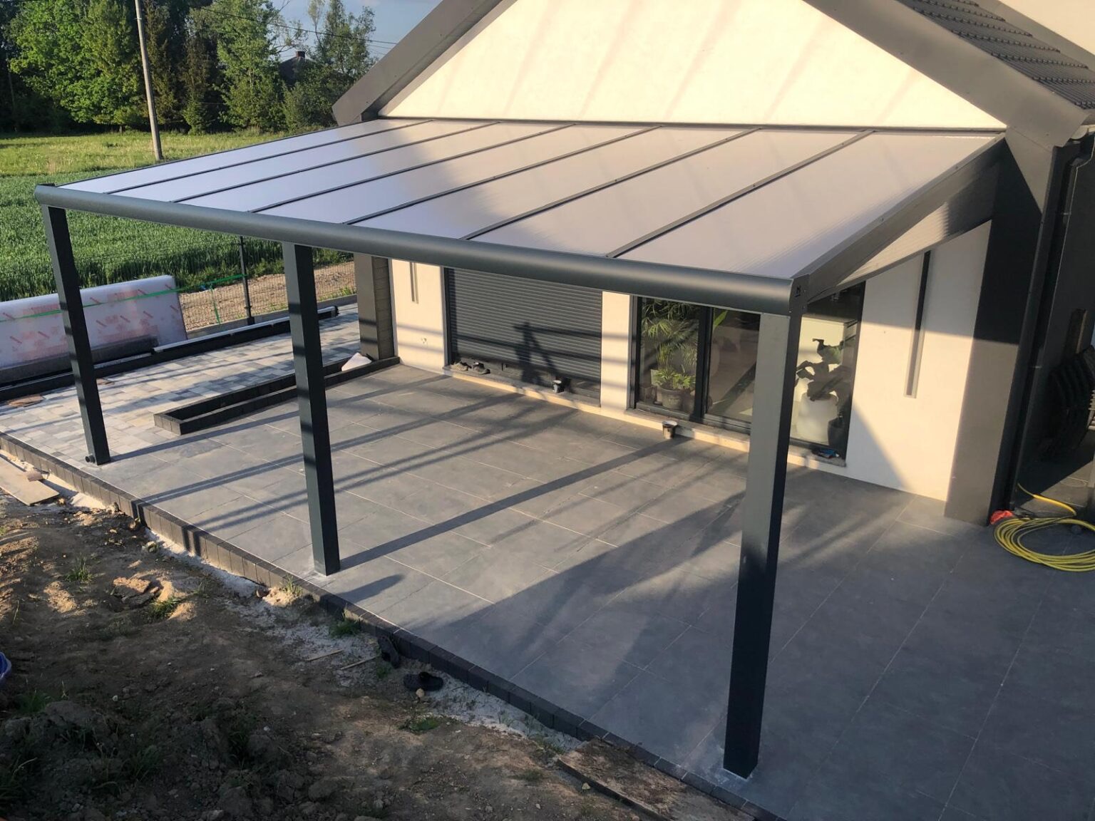 Nowoczesna, grafitowa pergola z poliwęglanu przy domu z szarym tarasem z płytek. Widok z dołu, w tle zieleń.