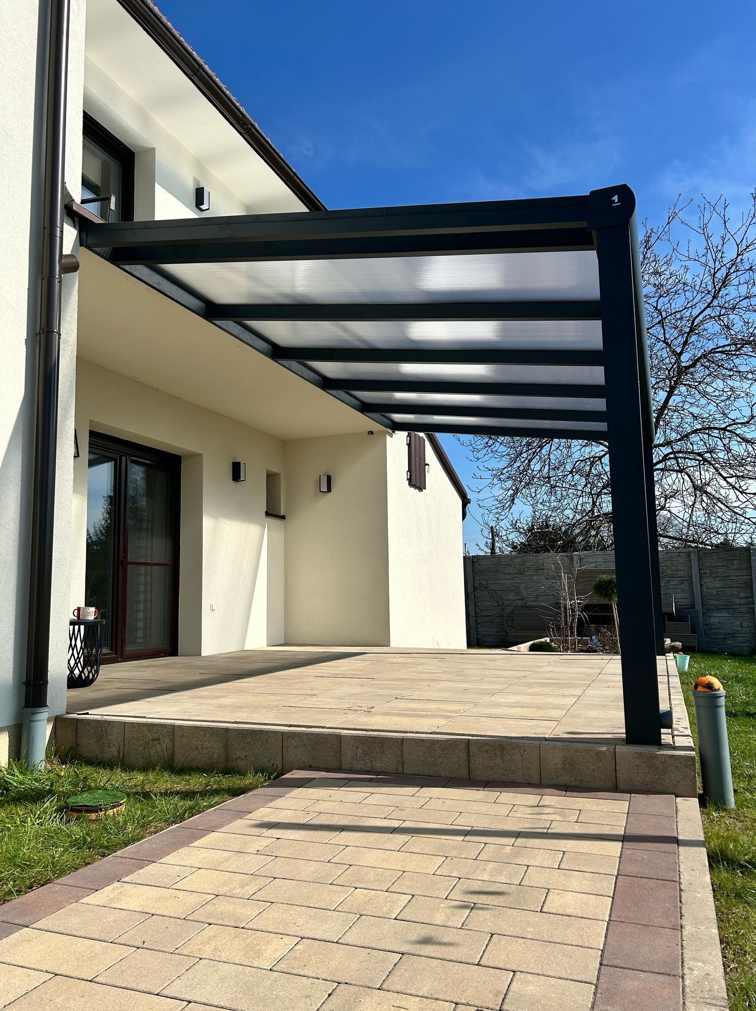 Nowoczesna, grafitowa pergola z przezroczystym zadaszeniem, przylegająca do jasnego budynku z brązowymi oknami. Widoczny taras z kostki brukowej i fragment ogrodu.