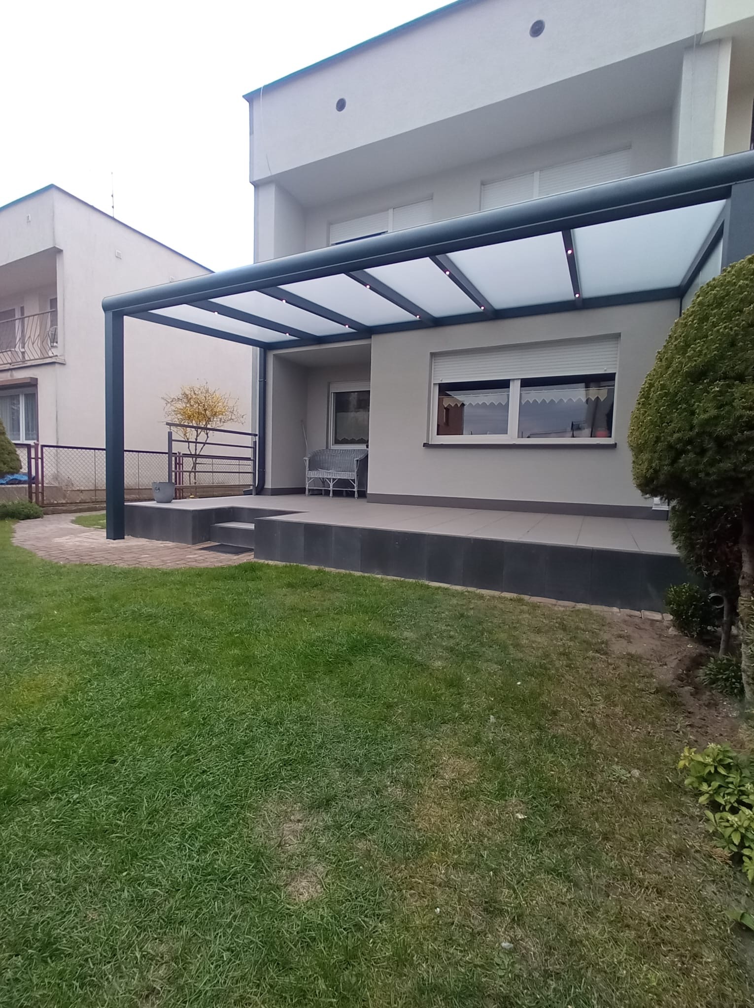 Nowoczesna pergola z szarym stelażem i mlecznym zadaszeniem, przylegająca do domu z tarasem. Widok z ogrodu, trawnik i elementy architektury ogrodowej.
