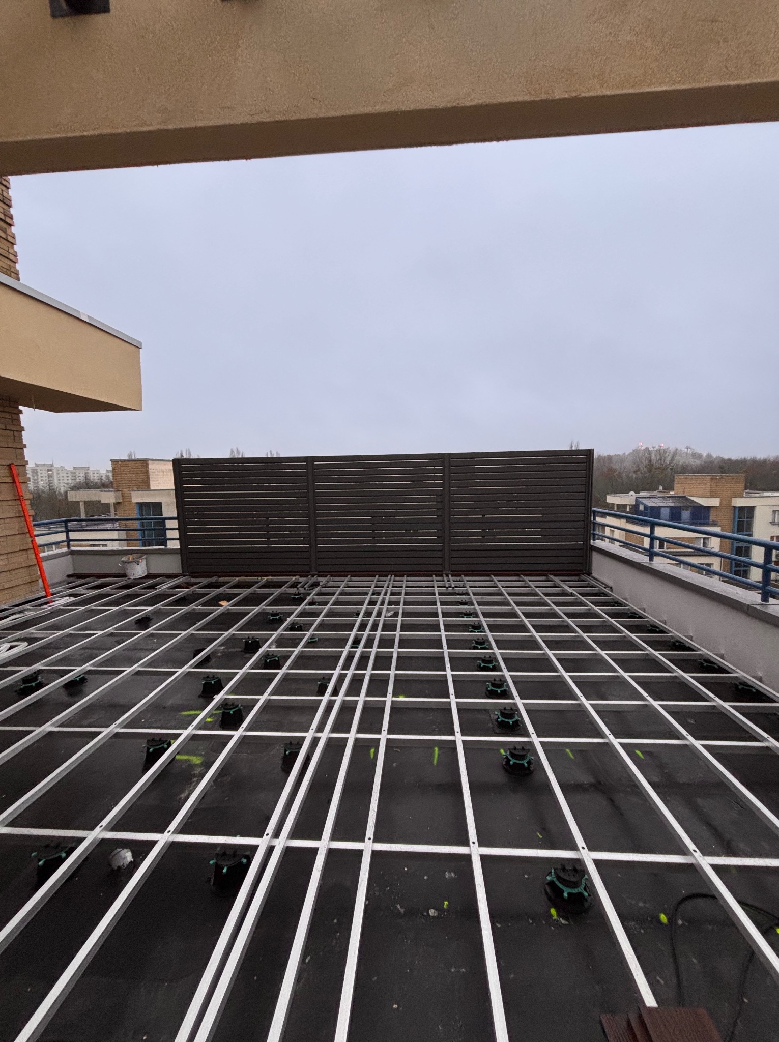 Montaż stelaża pod taras kompozytowy na balkonie. Widoczne profile aluminiowe i regulowane wsporniki. W tle zabudowa balkonu z poziomych desek.