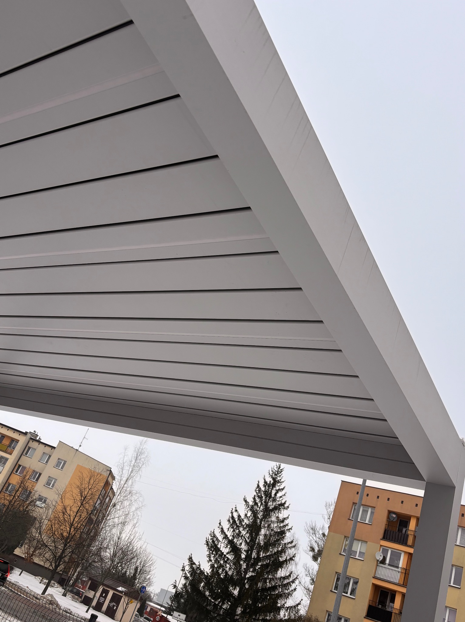 Nowoczesna, szara pergola z widokiem na budynki mieszkalne i drzewa w zimowej scenerii. Ujęcie z dołu, podkreślające minimalistyczny design konstrukcji.