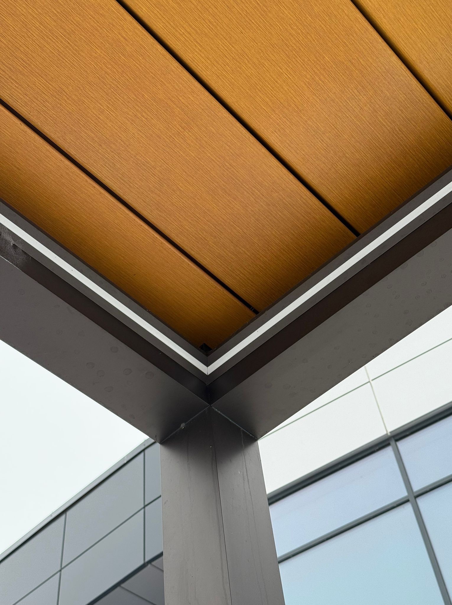 Nowoczesna pergola z brązowych desek kompozytowych, wsparta na ciemnoszarym słupie. Widok z dołu, z fragmentem budynku w tle.