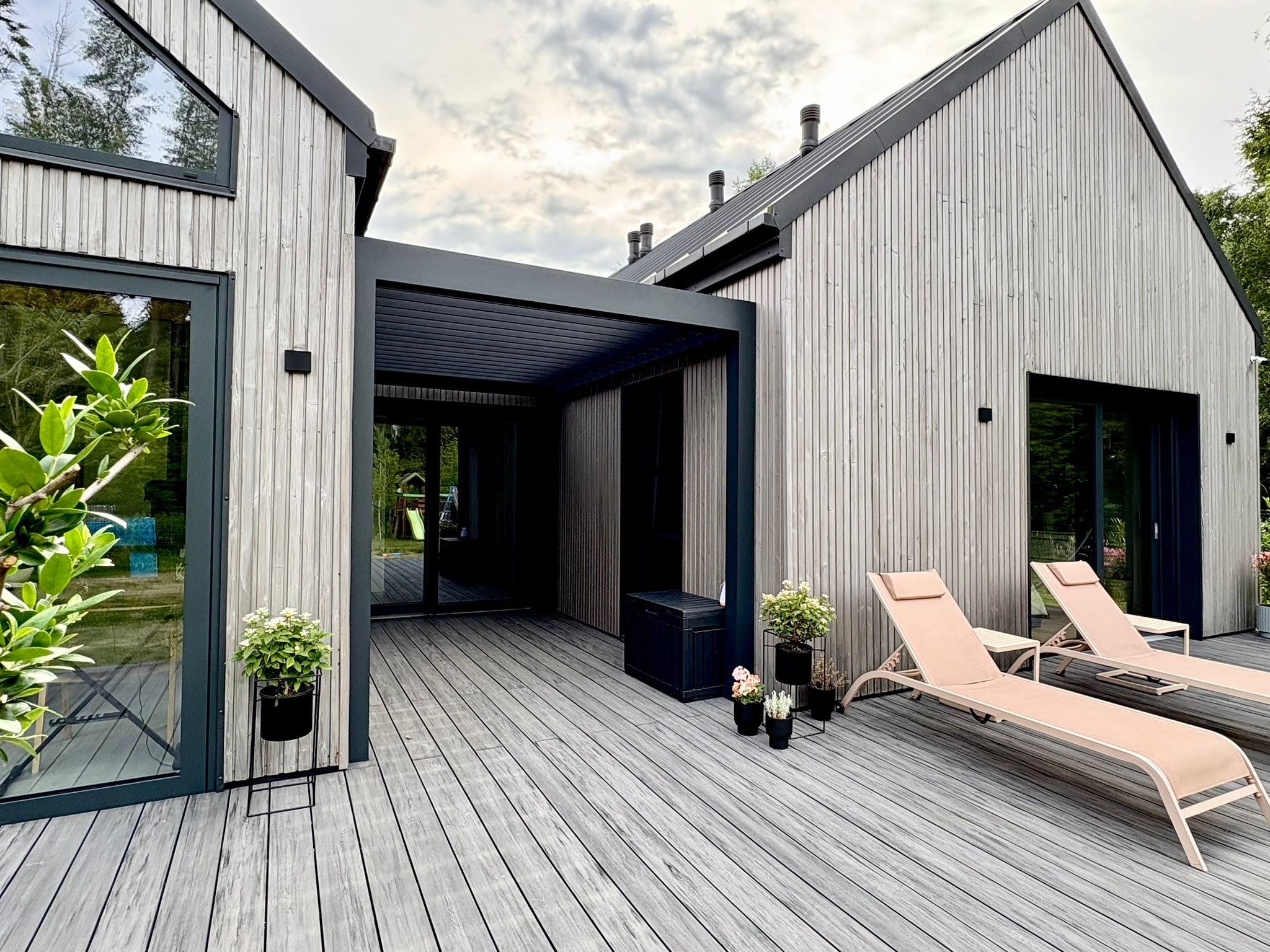 Nowoczesny taras kompozytowy z szarą deską, metalową pergolą i leżakami. Elewacja budynku z jasnego drewna. Minimalistyczny design, rośliny w donicach.