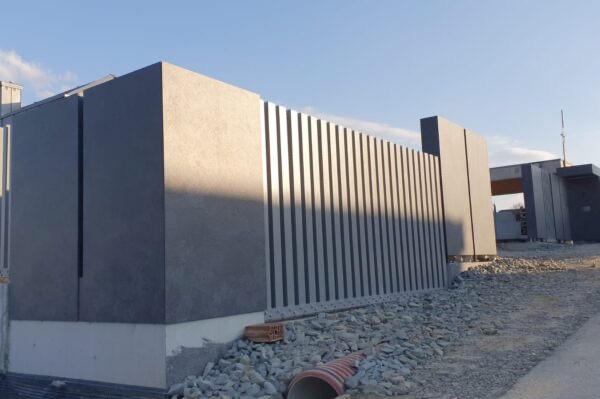 Nowoczesne ogrodzenie z szarych paneli pionowych i płyt, na fundamencie. Widoczne gruz i rura kanalizacyjna. Minimalistyczny design, surowy krajobraz.