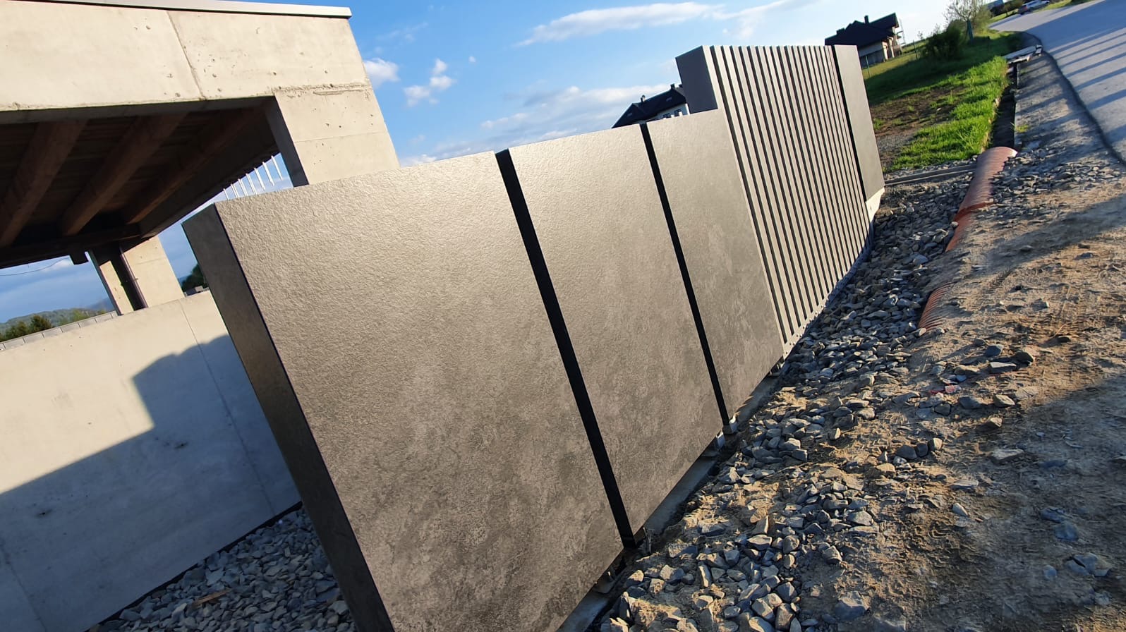 Nowoczesne ogrodzenie z paneli kompozytowych w odcieniach szarości, z widoczną strukturą i pionowymi żłobieniami, na tle błękitnego nieba i fragmentu betonowej konstrukcji.