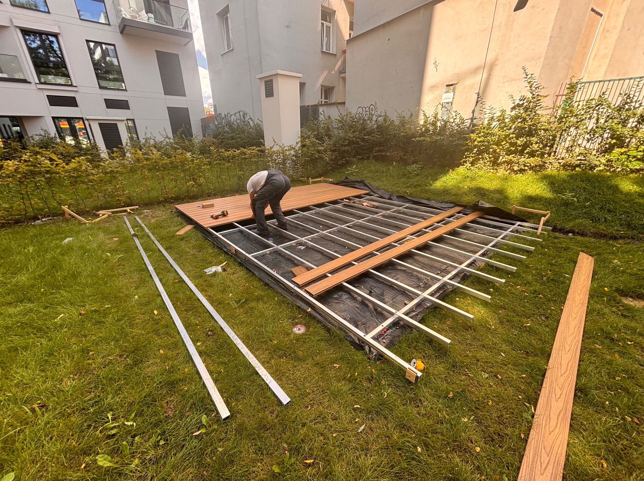 Montaż tarasu kompozytowego na trawie: pracownik układa deski na metalowej konstrukcji. Widoczne elementy mocujące i narzędzia. Tło stanowi budynek mieszkalny.
