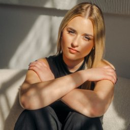 fredieme.photo - Alfred Wiśniewski - Portret młodej kobiety z blond włosami, siedzącej na sofie w kontraście światła i cienia, z rękami skrzyżowanymi na piersiach, w czarnym stroju.