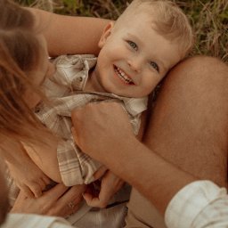 fredieme.photo - Alfred Wiśniewski - Uśmiechnięty blond chłopiec w ramionach rodziców, leżący na trawie. Ciepłe, naturalne światło podkreśla bliskość i radość rodzinnej sesji plenerowej.