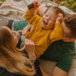 fredieme.photo - Alfred Wiśniewski - Radosny chłopiec w żółtej bluzie, trzymający czerwoną zabawkę, leży na kocu z rodzicami. Tata całuje synka w głowę, mama patrzy z uśmiechem. Jesienne liście w tle.