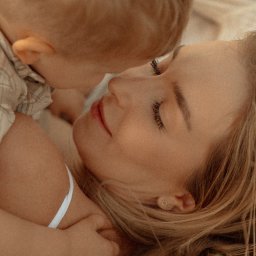 fredieme.photo - Alfred Wiśniewski - Delikatne zbliżenie: blondwłosa mama z zamkniętymi oczami, przytulona do małego dziecka w beżowej koszuli, subtelne światło, naturalna sceneria, intymna chwila.