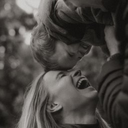 fredieme.photo - Alfred Wiśniewski - Czarno-białe zdjęcie matki śmiejącej się, trzymającej w górze małe dziecko, które również się śmieje, z widocznym fragmentem sylwetki ojca w tle.