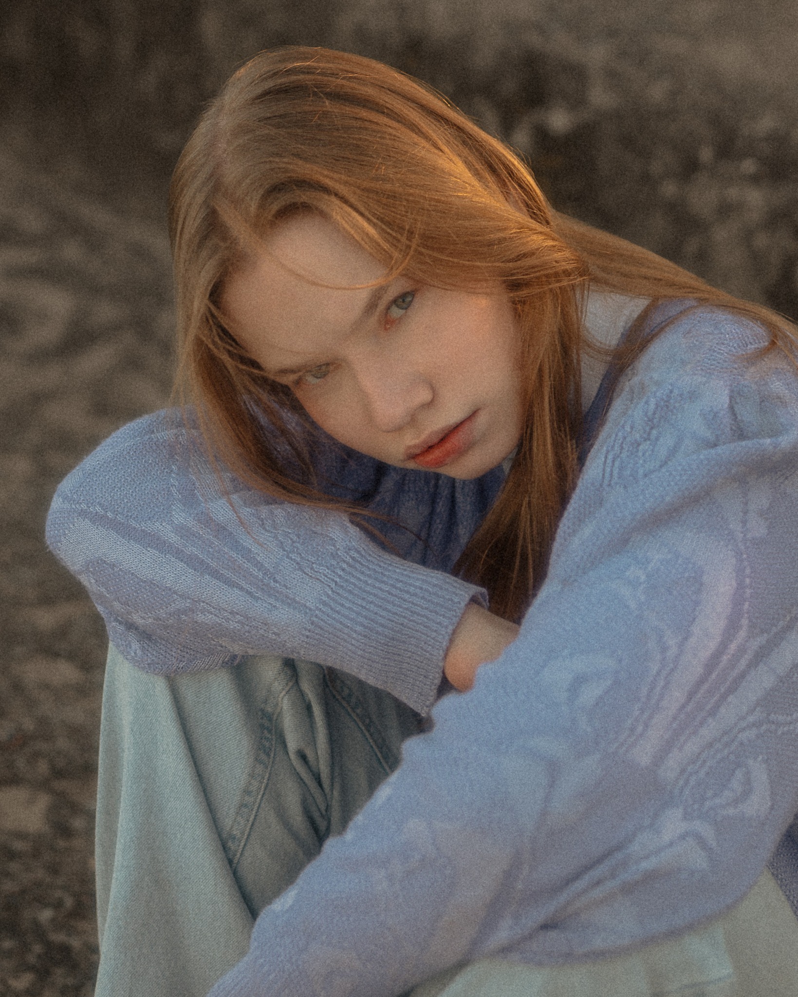 Portret młodej kobiety o rudych włosach, ubranej w błękitny sweter, siedzącej na tle szarej, teksturowanej powierzchni. Delikatne światło podkreśla rysy twarzy.