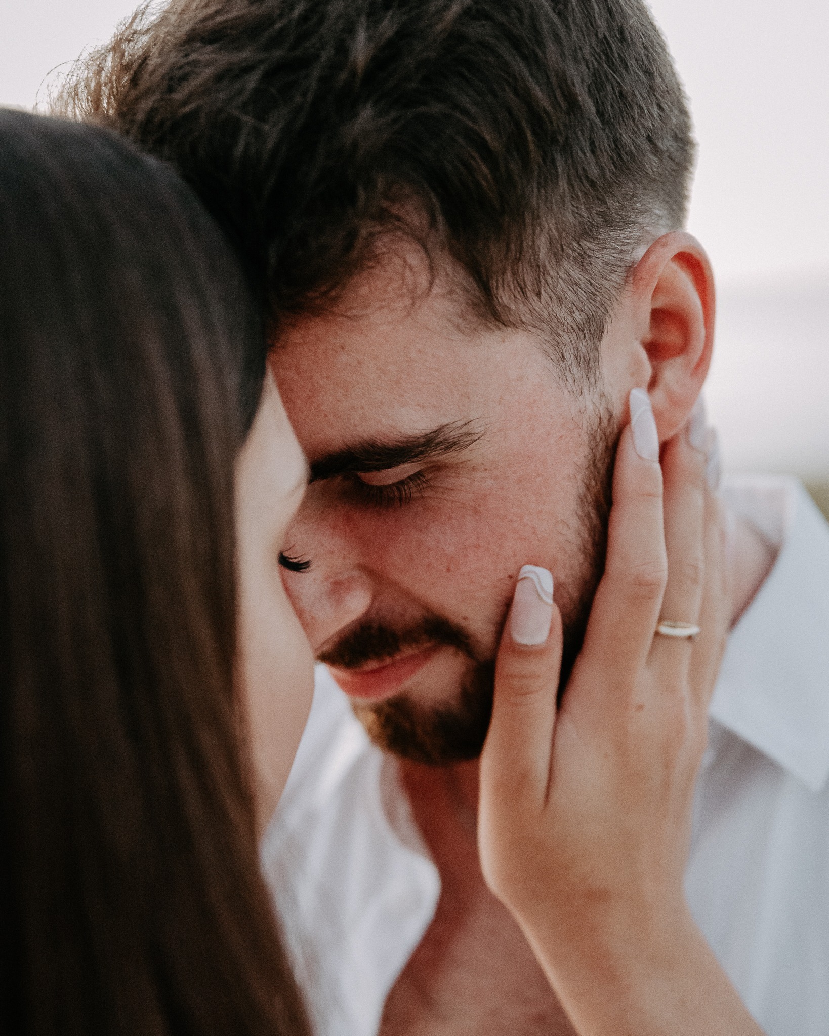 Zbliżenie na twarz mężczyzny z brodą, dotykaną przez kobietę z pierścionkiem. Delikatne światło podkreśla intymność chwili. Romantyczna sesja w plenerze.