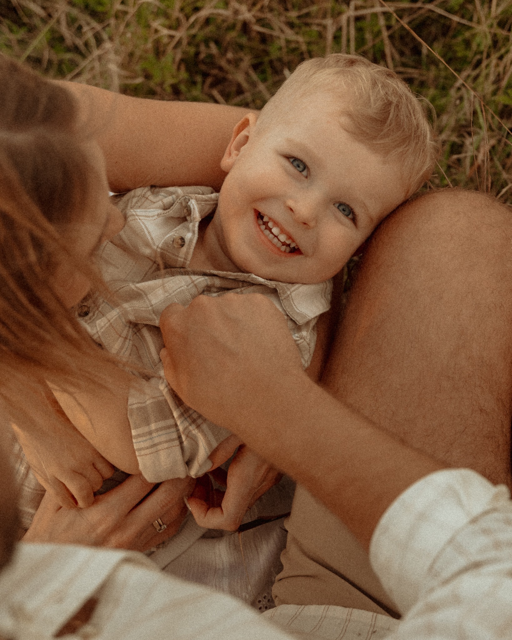 Uśmiechnięty blond chłopiec w ramionach rodziców, leżący na trawie. Ciepłe, naturalne światło podkreśla bliskość i radość rodzinnej sesji plenerowej.