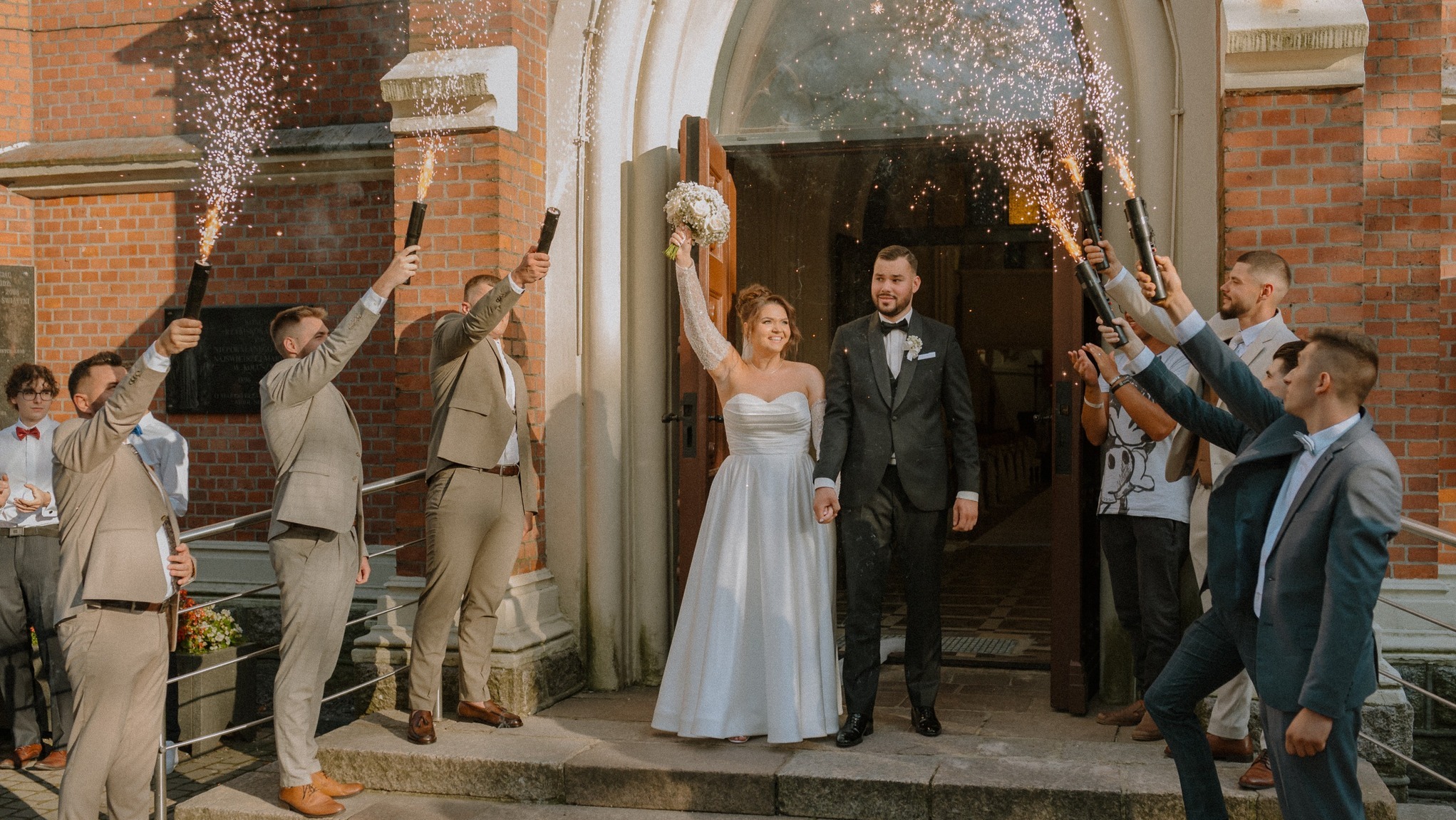 Para młoda wychodzi z kościoła, otoczona przez gości z racami. Panna młoda w białej sukni, pan młody w garniturze. Ceglany budynek kościoła w tle, ciepłe światło.