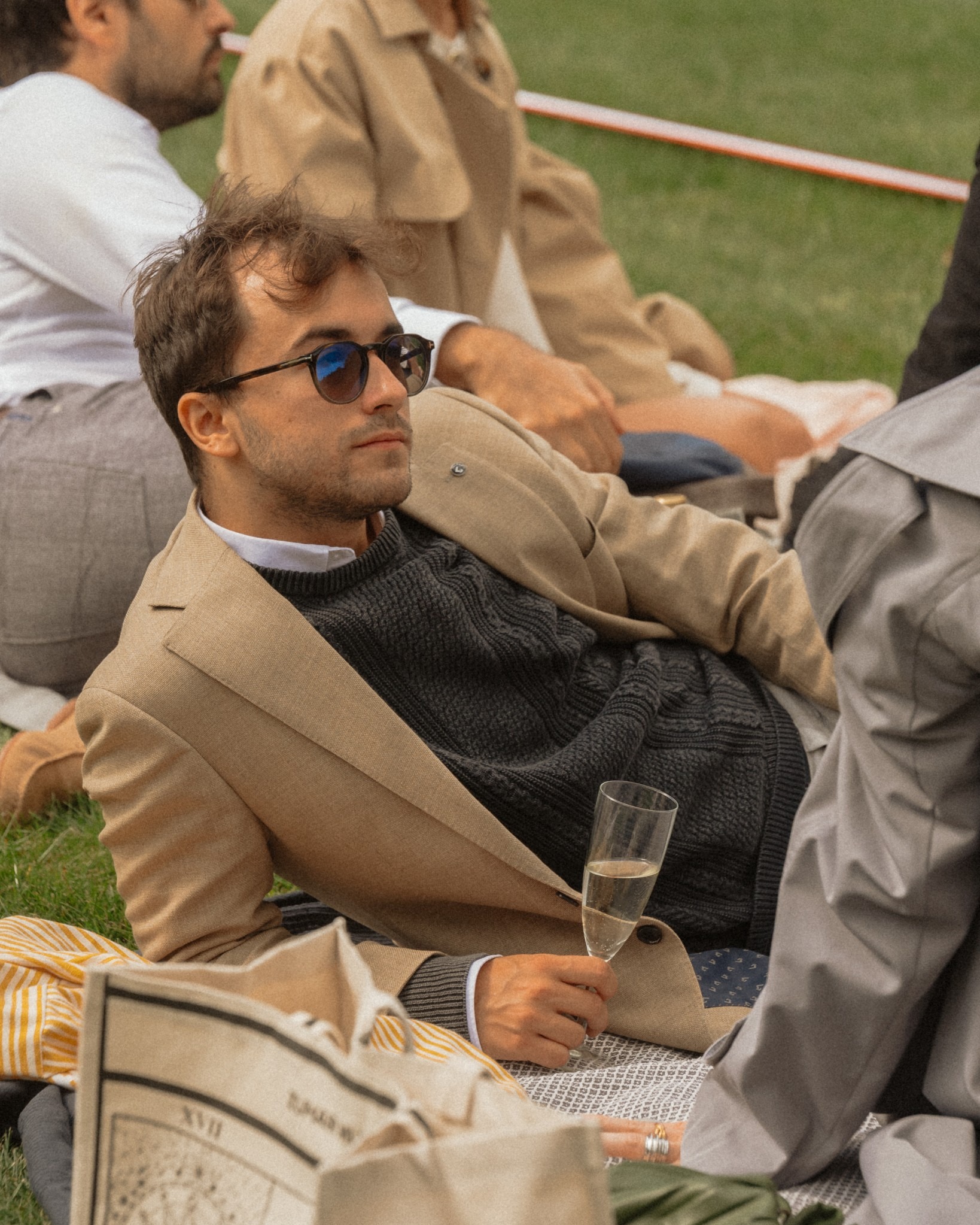 Elegancki mężczyzna w okularach przeciwsłonecznych relaksuje się na pikniku z kieliszkiem szampana, otoczony przez innych ludzi w stylowych ubraniach na trawie.