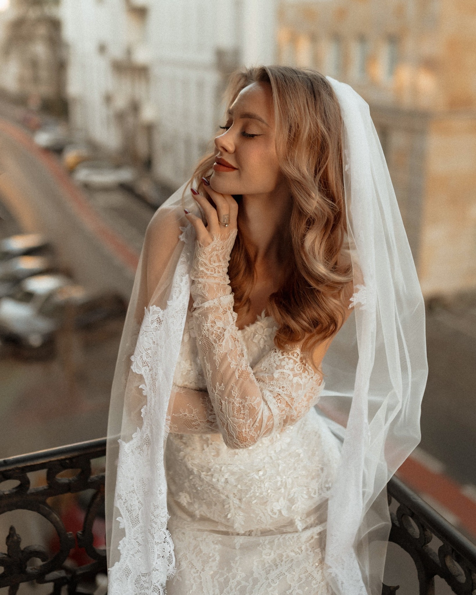 Panna młoda w koronkowej sukni i welonie, z zamkniętymi oczami, pozuje na balkonie z widokiem na miasto w tle, w ciepłym, naturalnym oświetleniu.