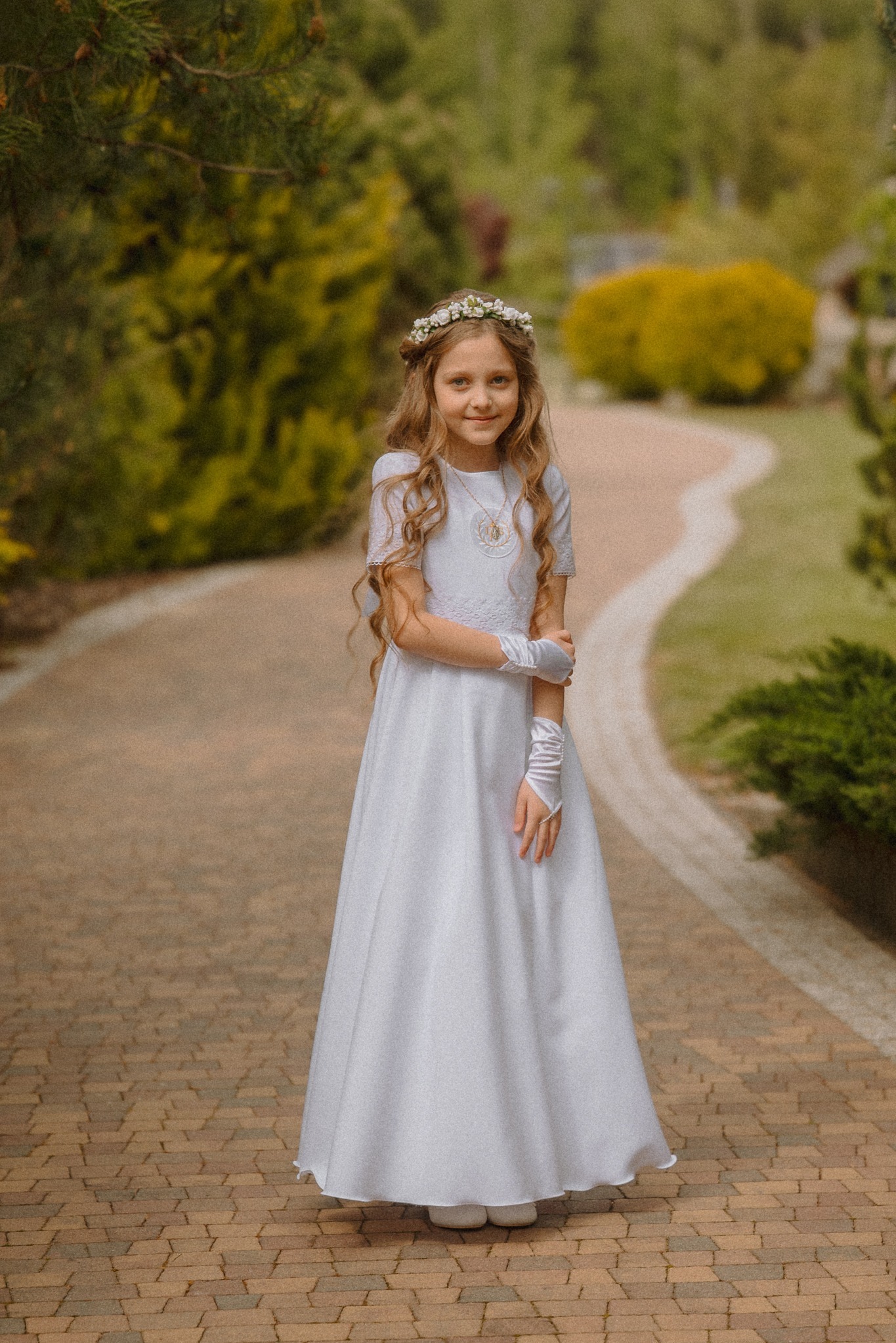Dziewczynka w białej sukience komunijnej, z wiankiem na głowie, pozuje na ceglanej ścieżce w ogrodzie. Uśmiechnięta, z delikatną biżuterią i rękawiczkami.
