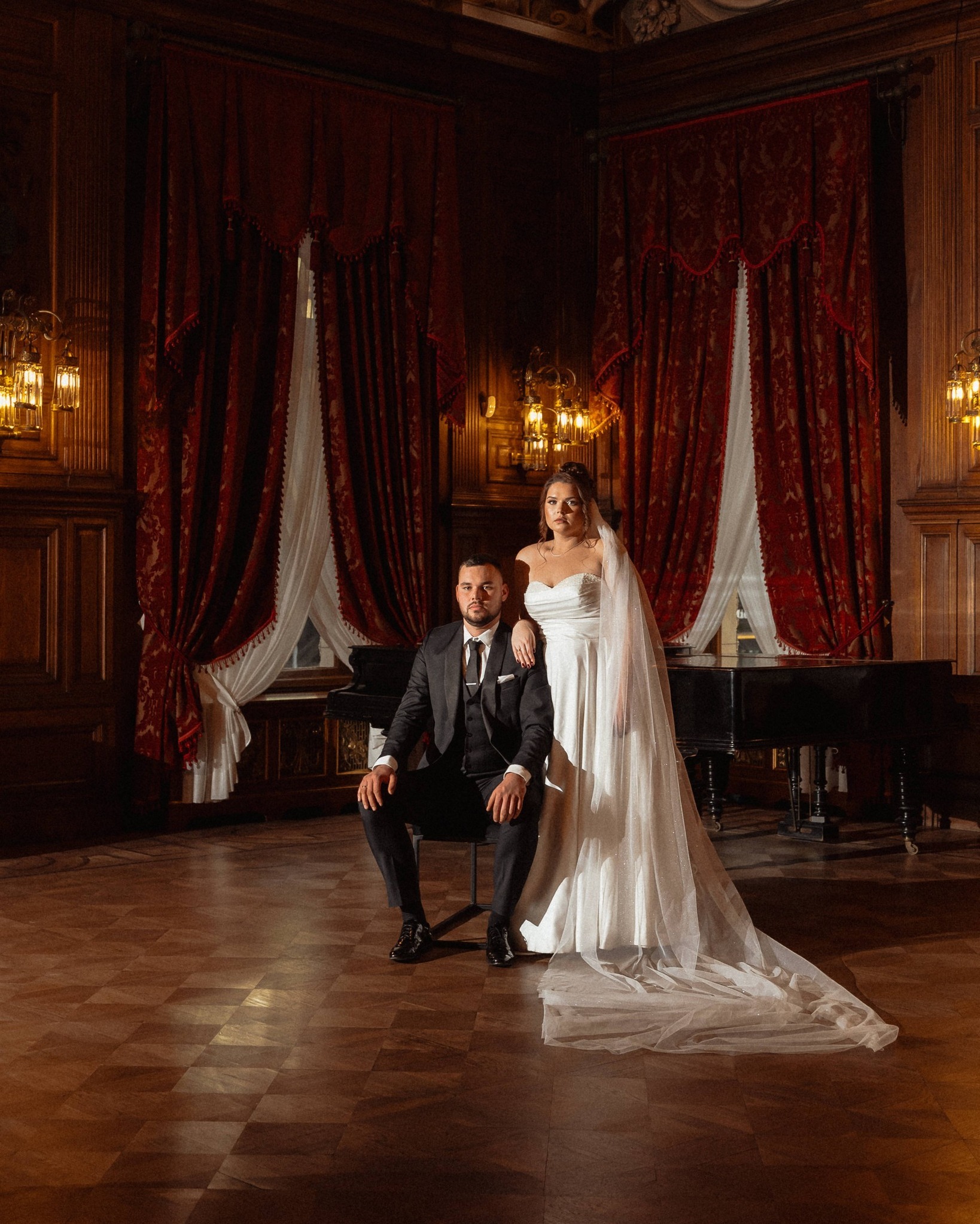 Elegancka sesja ślubna w pałacowym wnętrzu: para młoda pozuje na tle fortepianu i czerwonych kotar. Pan młody siedzi, panna młoda stoi obok.
