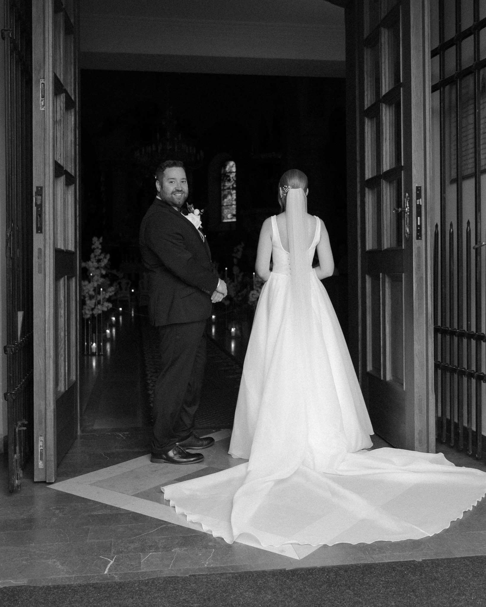 Czarno-białe zdjęcie pary młodej stojącej w drzwiach kościoła. Pan młody w garniturze, panna młoda w sukni z długim trenem. W tle widoczne świece i witraże.