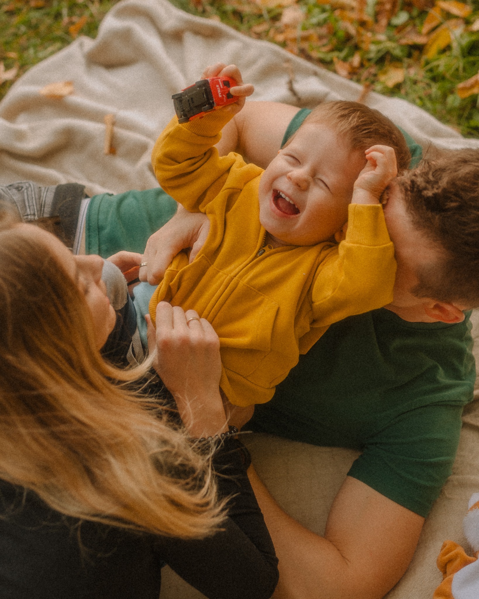 Radosny chłopiec w żółtej bluzie, trzymający czerwoną zabawkę, leży na kocu z rodzicami. Tata całuje synka w głowę, mama patrzy z uśmiechem. Jesienne liście w tle.