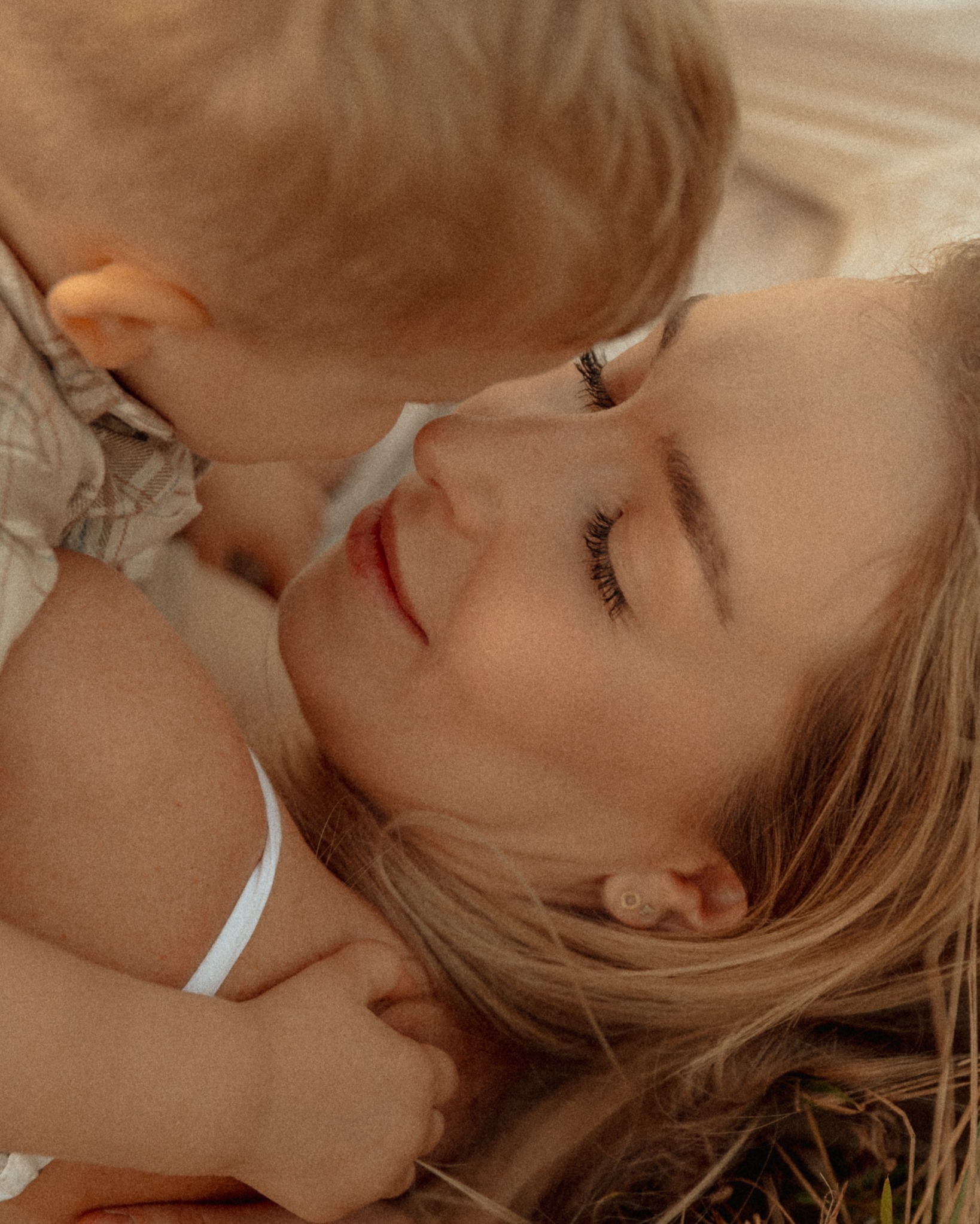Delikatne zbliżenie: blondwłosa mama z zamkniętymi oczami, przytulona do małego dziecka w beżowej koszuli, subtelne światło, naturalna sceneria, intymna chwila.