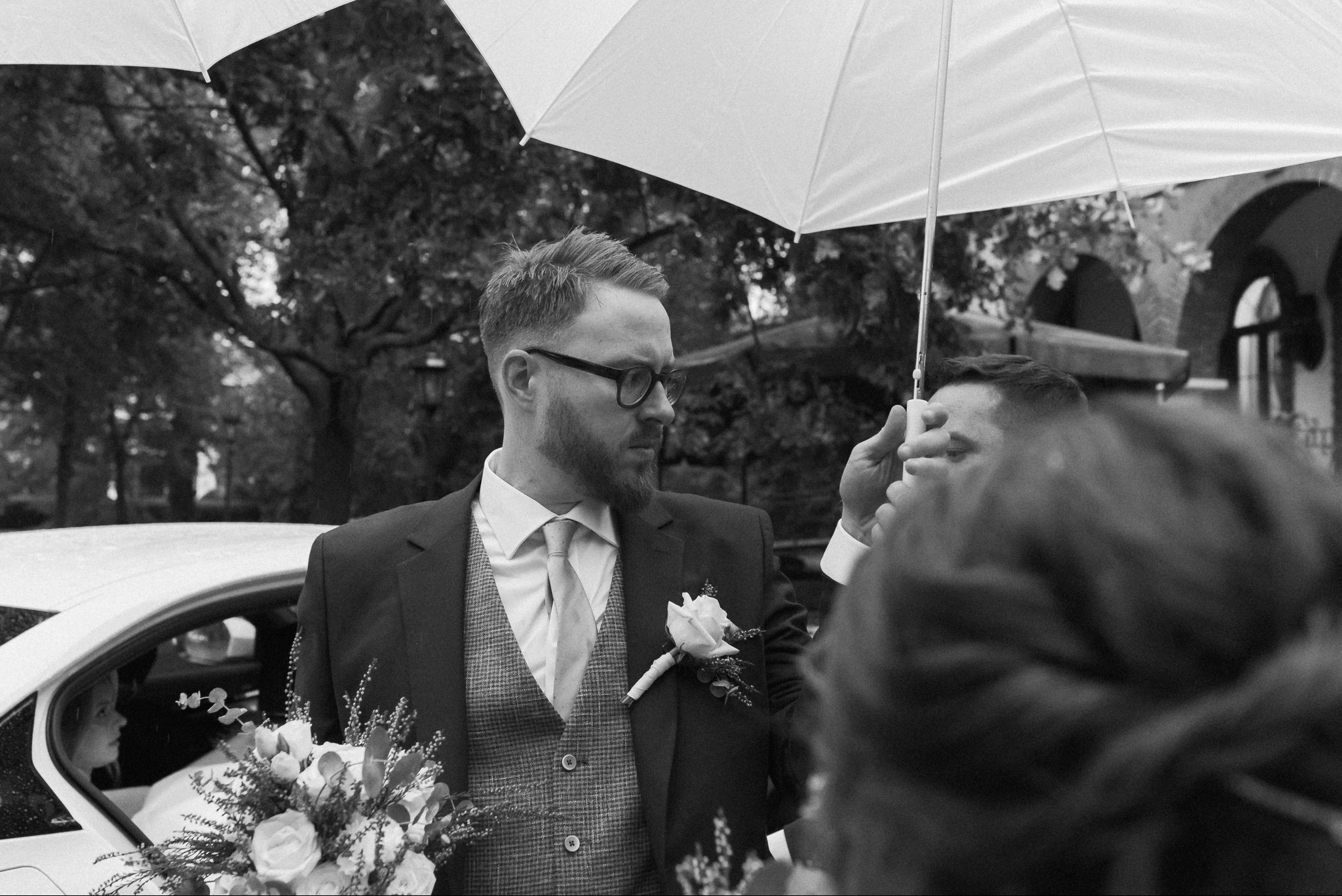 Czarno-białe zdjęcie pana młodego w garniturze, z bukietem i butonierką, pod parasolem, zarys samochodu i osoby w tle. Ujęcie z reportażu ślubnego.