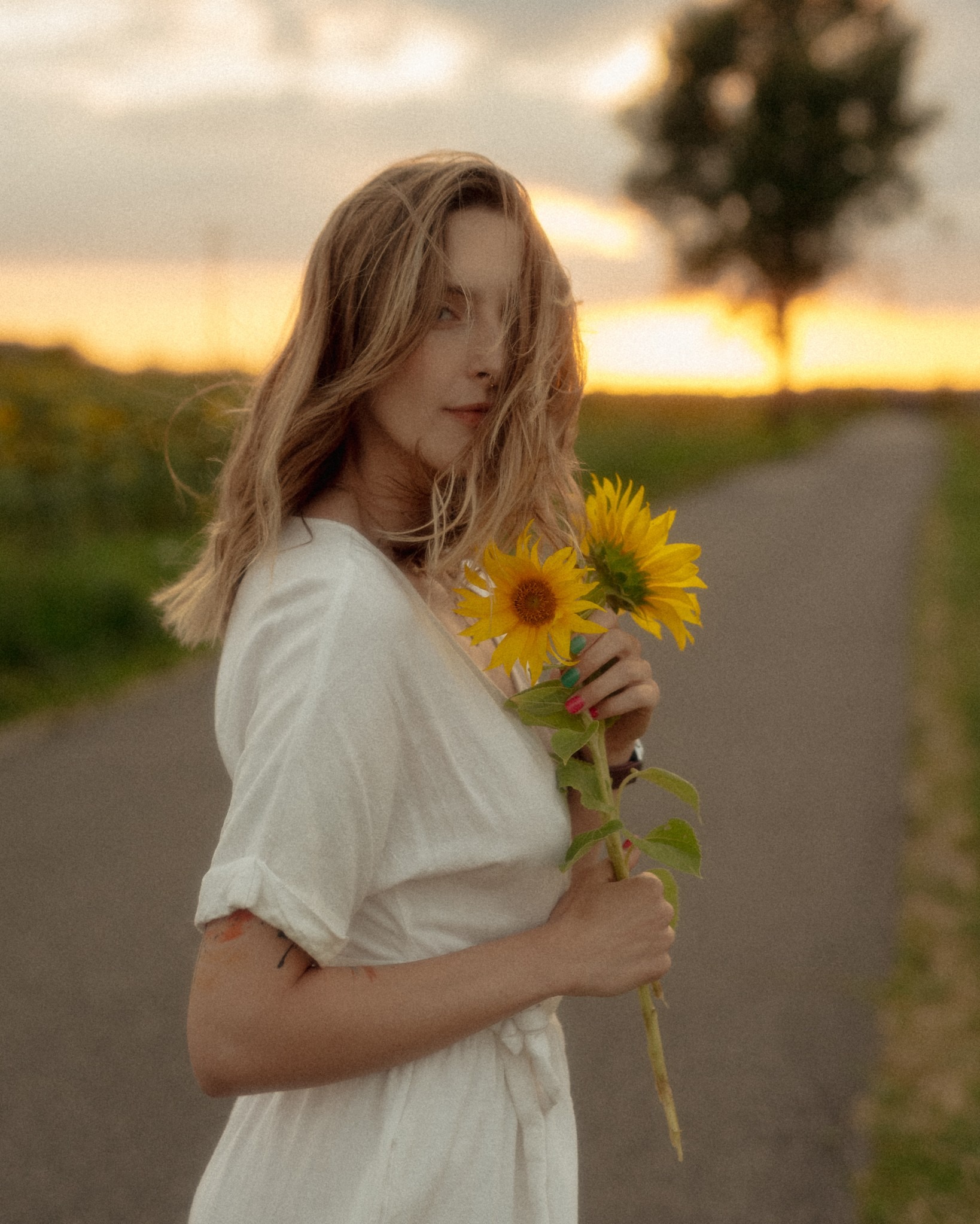Portret kobiety w białej sukience trzymającej dwa słoneczniki na tle drogi i zachodzącego słońca. Delikatne, naturalne światło podkreśla subtelność ujęcia.