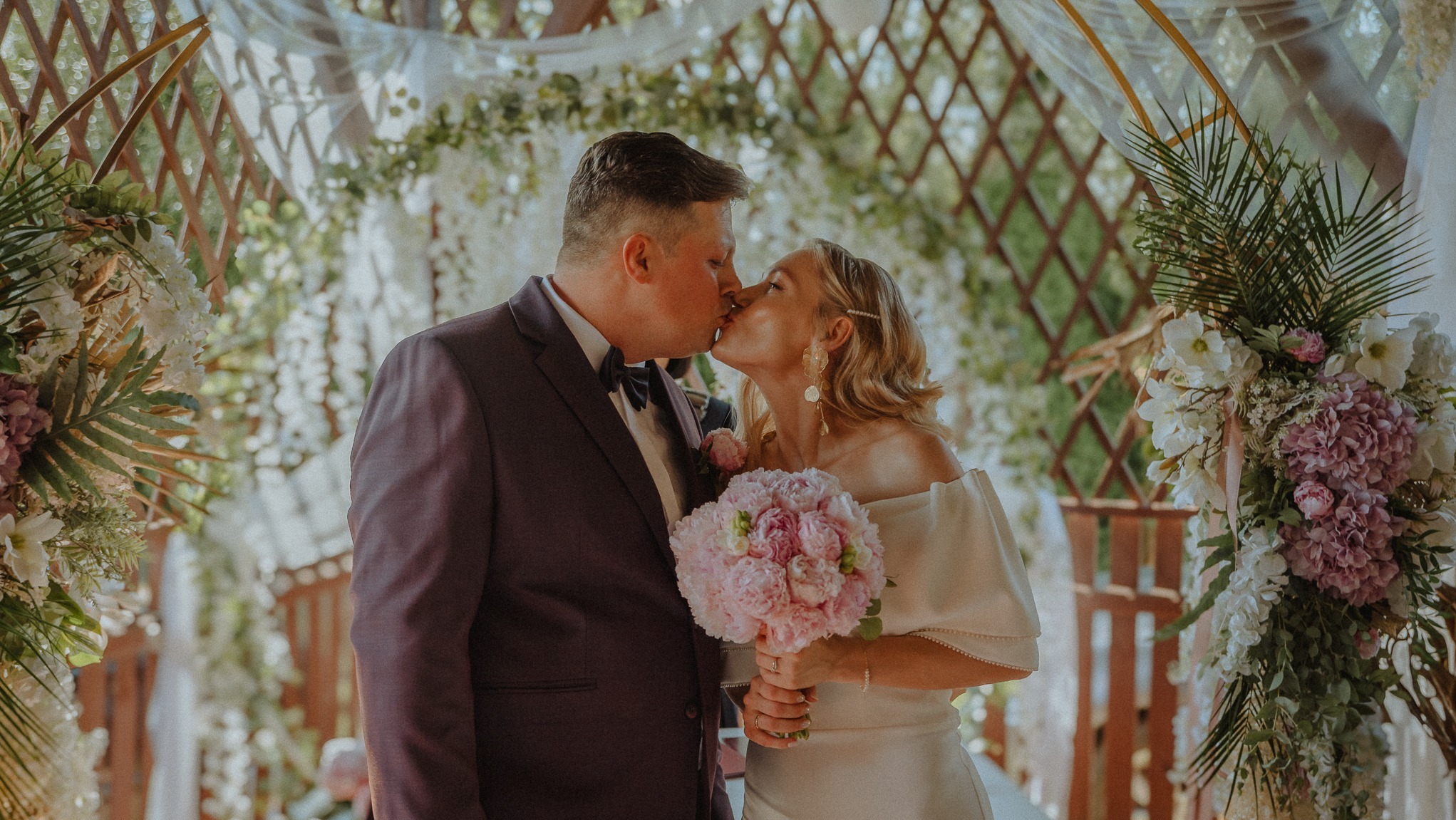 Pocałunek pary młodej pod drewnianą altaną udekorowaną kwiatami w odcieniach bieli i różu. Panna młoda trzyma bukiet z piwonii.