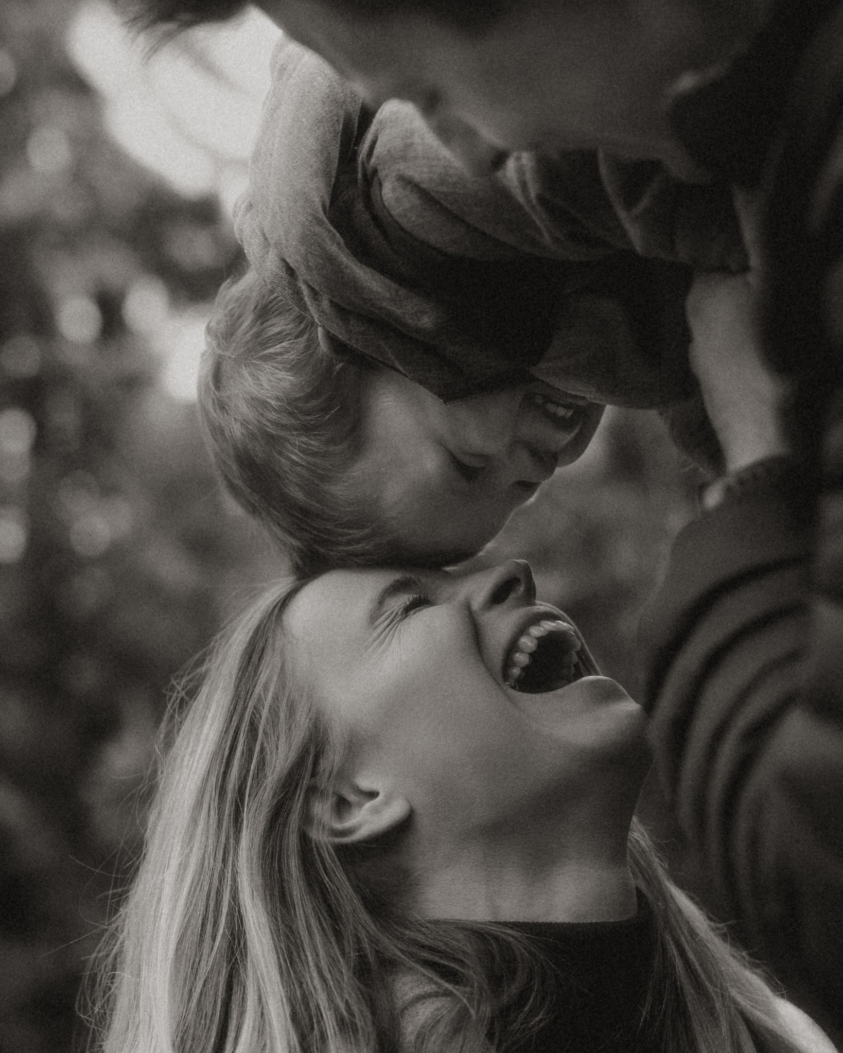 Czarno-białe zdjęcie matki śmiejącej się, trzymającej w górze małe dziecko, które również się śmieje, z widocznym fragmentem sylwetki ojca w tle.