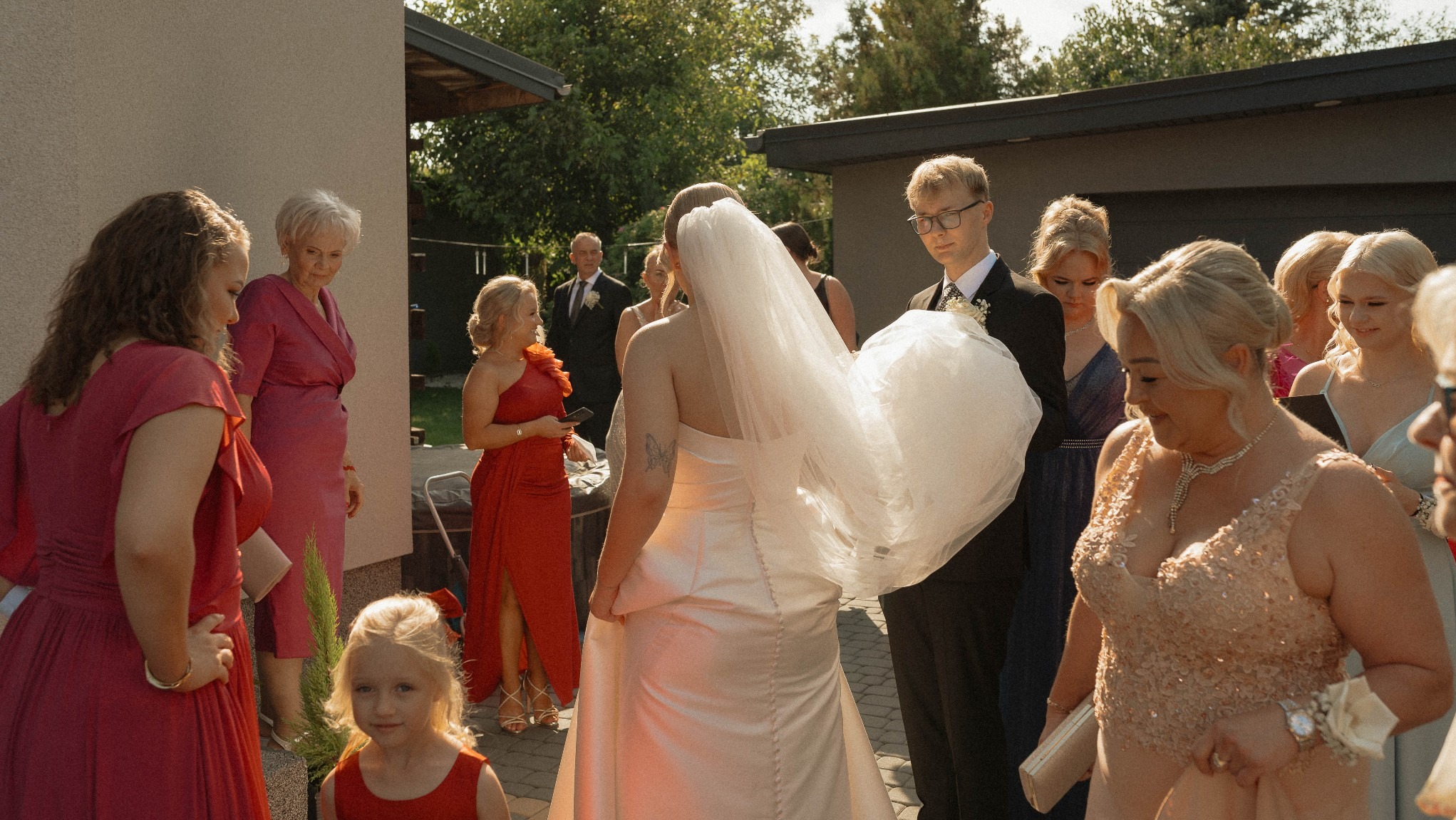 Panna młoda w sukni ślubnej z welonem, otoczona gośćmi weselnymi w ogrodzie, uchwycona z perspektywy pleców, z widocznym tatuażem motyla.