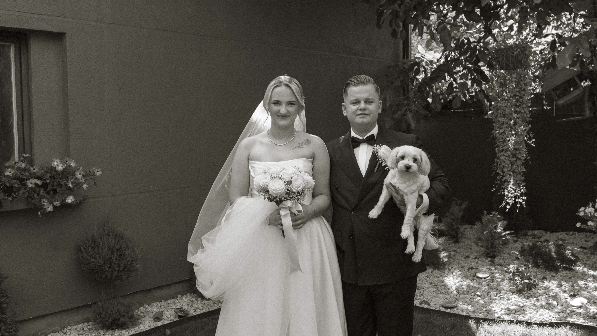 Czarno-białe zdjęcie pary młodej z psem rasy Maltańczyk na zewnątrz, panna młoda trzyma bukiet białych róż, a pan młody ma muchę i kwiat w butonierce.