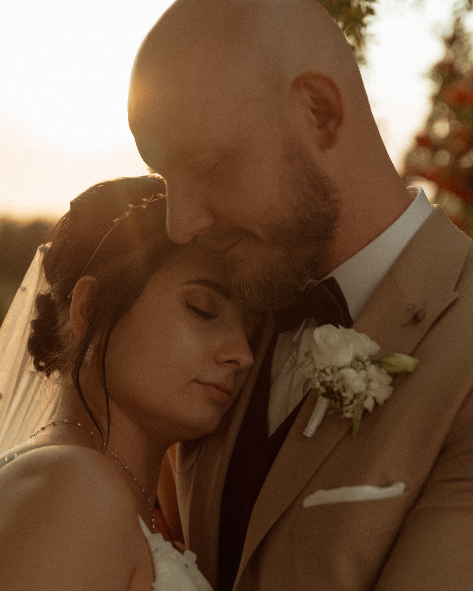 Delikatne ujęcie pary młodej w objęciach, w ciepłym świetle zachodzącego słońca. Panna młoda z zamkniętymi oczami opiera się o ramię pana młodego w beżowym garniturze.