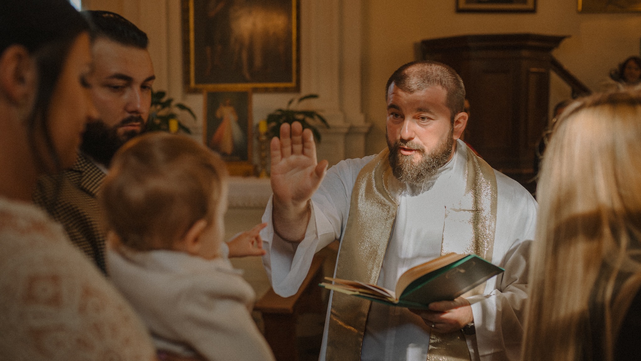 Ksiądz z brodą błogosławi dziecko podczas ceremonii chrztu w kościele, w tle rodzina i obrazy religijne. Ciepłe oświetlenie podkreśla uroczysty charakter.