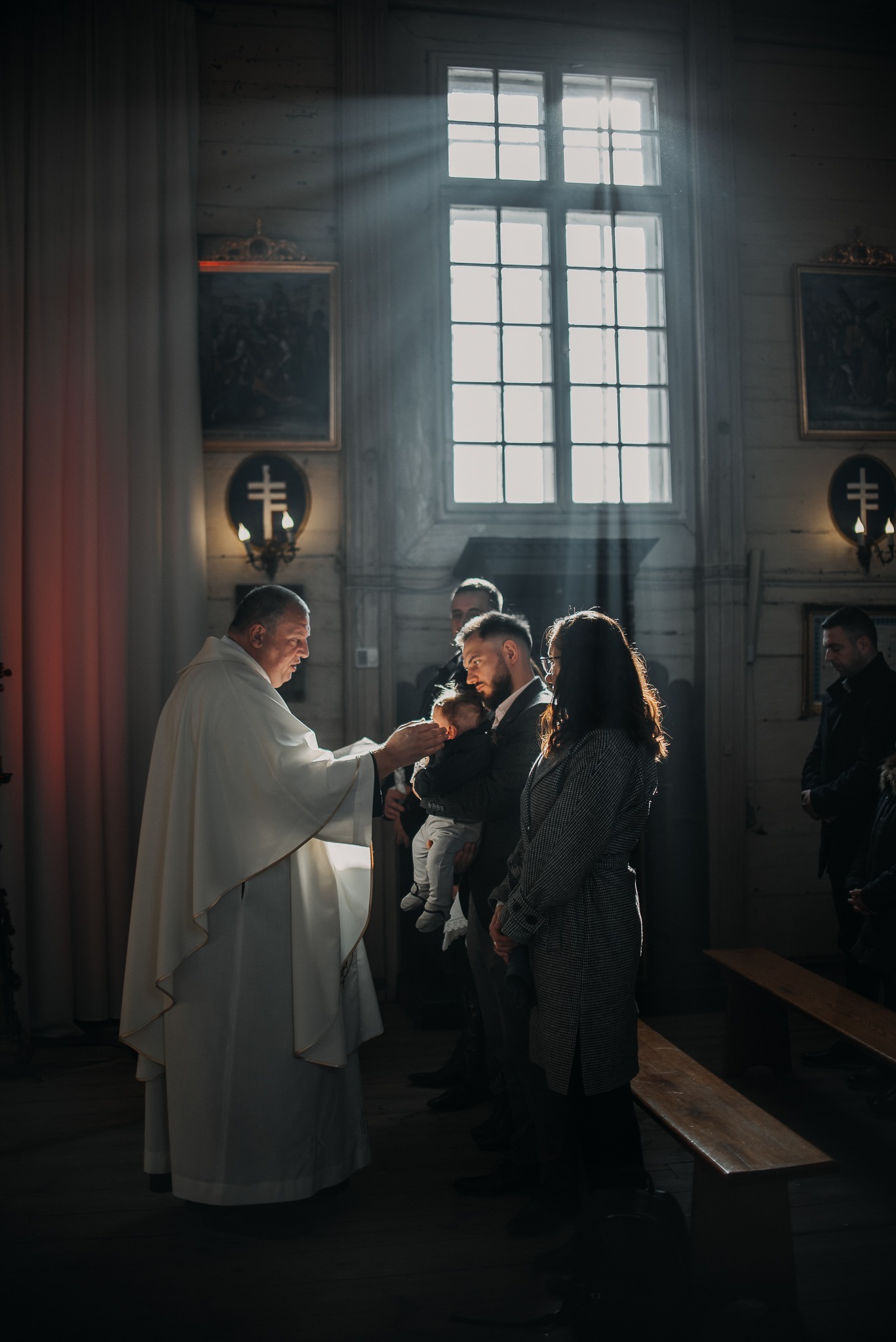 Ksiądz udziela sakramentu chrztu dziecku w kościele, w tle rodzina i promienie światła wpadające przez okno. Uroczysta atmosfera, drewniane wnętrze.