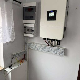 Sigma Services SZYMON GRUŻALSKI - Biały inwerter fotowoltaiczny i akumulator energii zamontowane na ścianie wewnątrz budynku, widoczne kable i elementy instalacji elektrycznej.