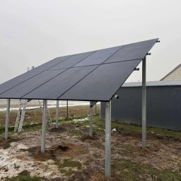 Sigma Services SZYMON GRUŻALSKI - Montaż paneli fotowoltaicznych na konstrukcji wsporczej w Spławiu. Widoczna drabina i przygotowany teren pod instalację. Pochmurne niebo w tle.