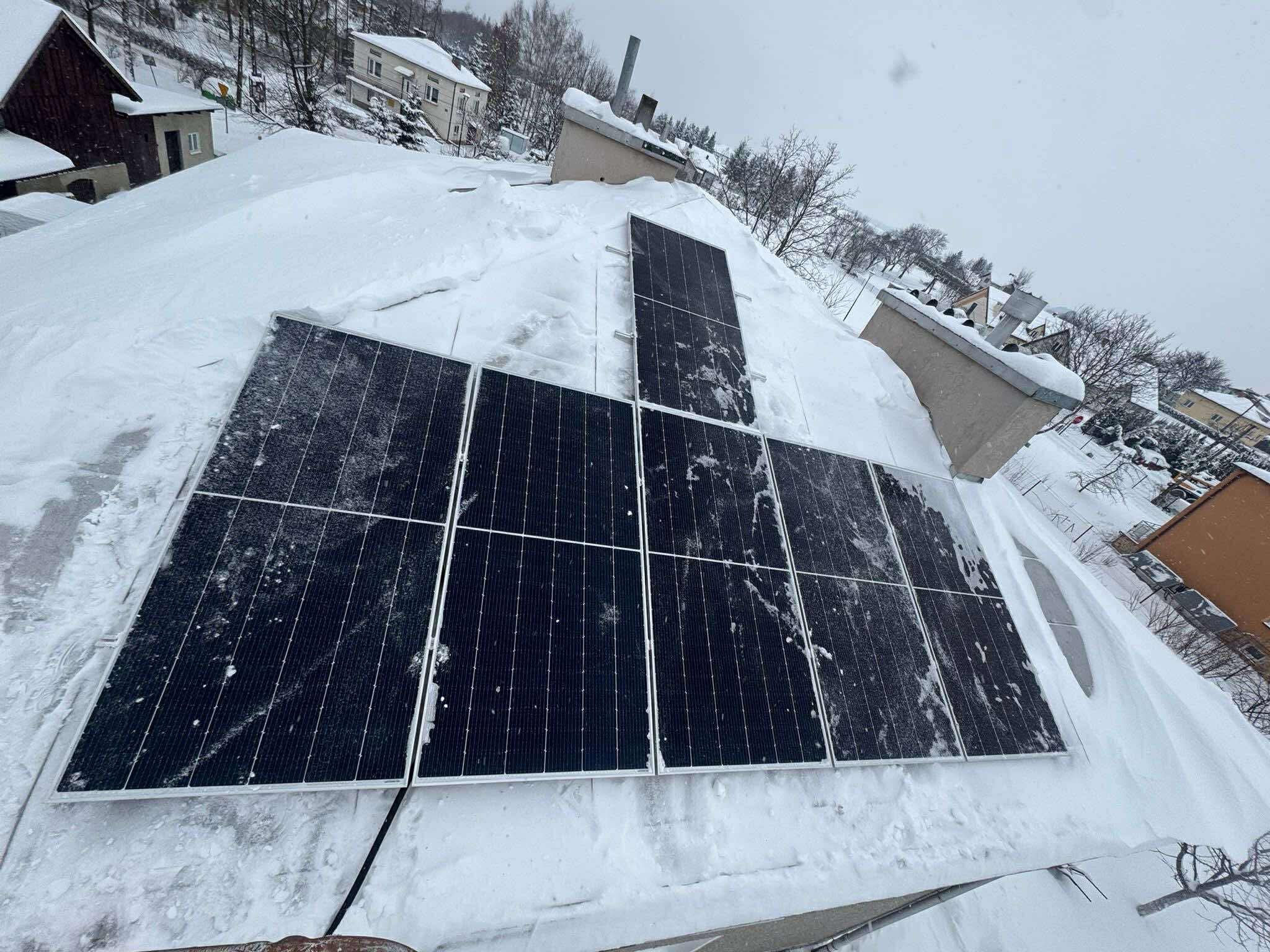 Panele fotowoltaiczne pokryte śniegiem na dachu budynku w zimowej scenerii. Widok z góry, pochmurne niebo, zabudowa w tle. Częściowo odśnieżone moduły.
