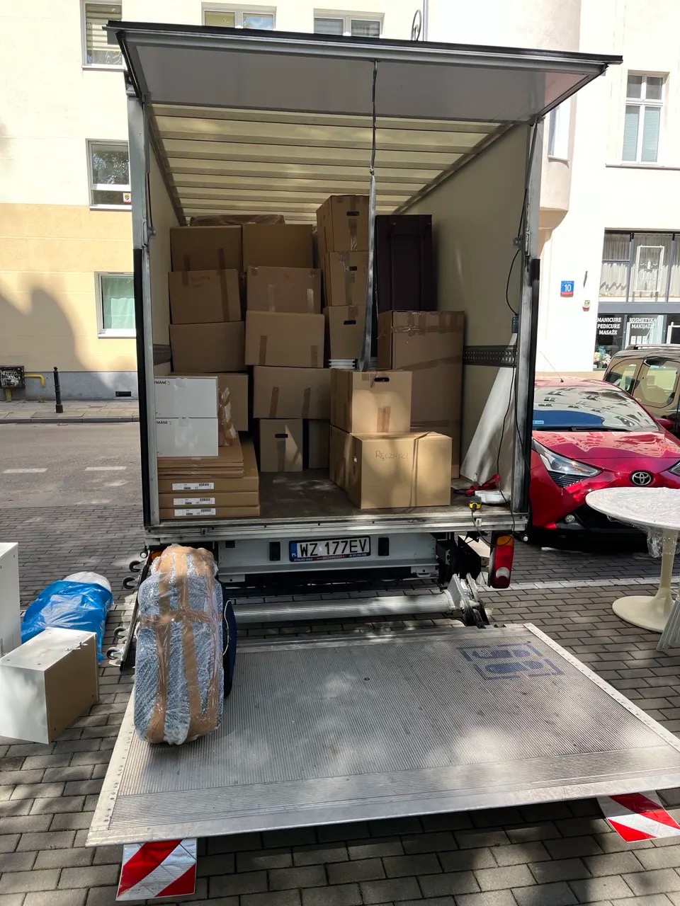 Otwarty bus transportowy wypełniony kartonami i zabezpieczonymi meblami, gotowy do transportu na ulicy miejskiej w słoneczny dzień. Widoczny numer rejestracyjny.